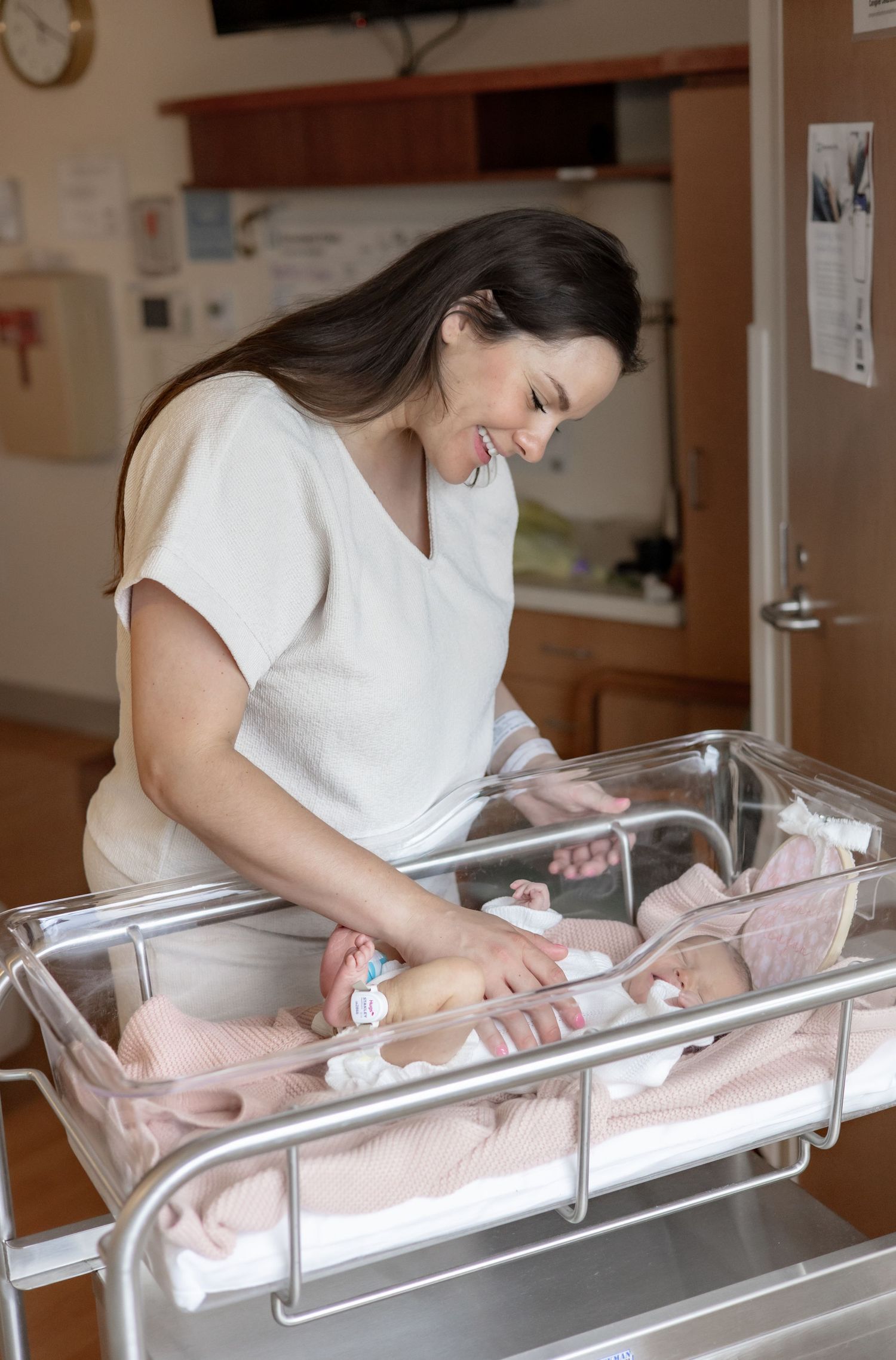 A healthcare worker in a white shirt smiles while tending to a newborn baby in a hospital bassinet.