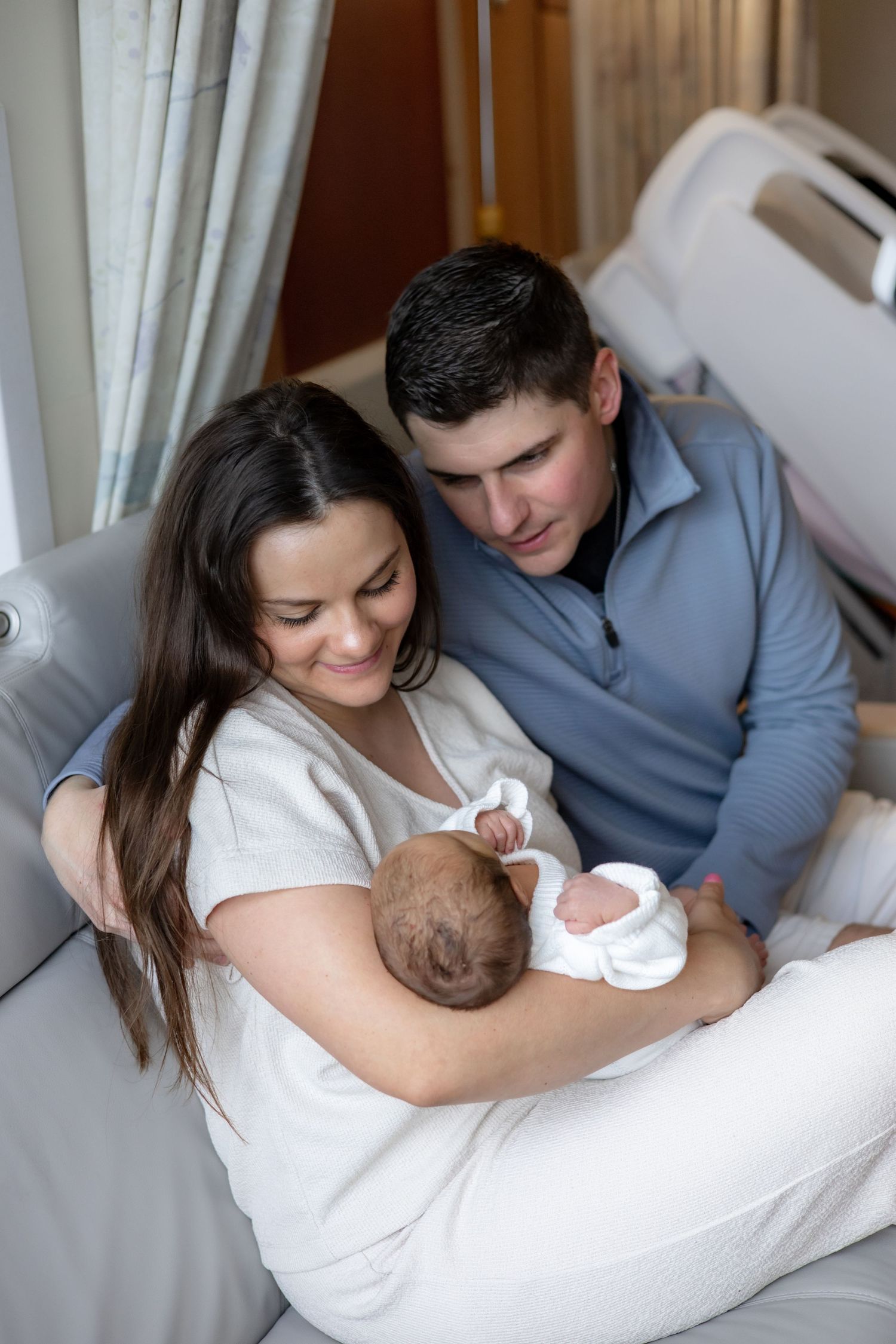Parents sharing intimate bonding moments with their newborn baby while resting together on a white bed.