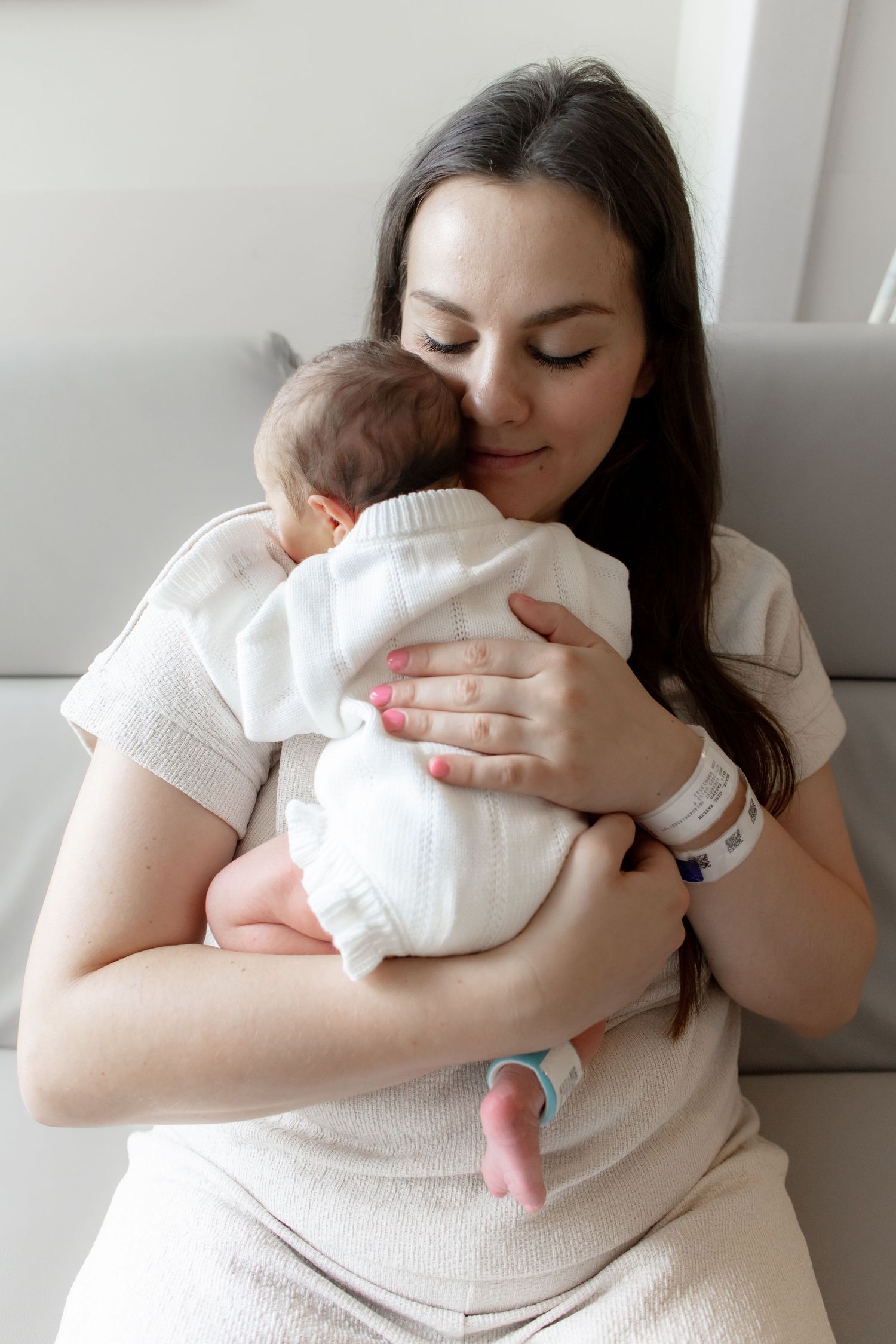 A tender moment as a mother in white clothing cuddles her sleeping newborn baby wrapped in a white blanket.