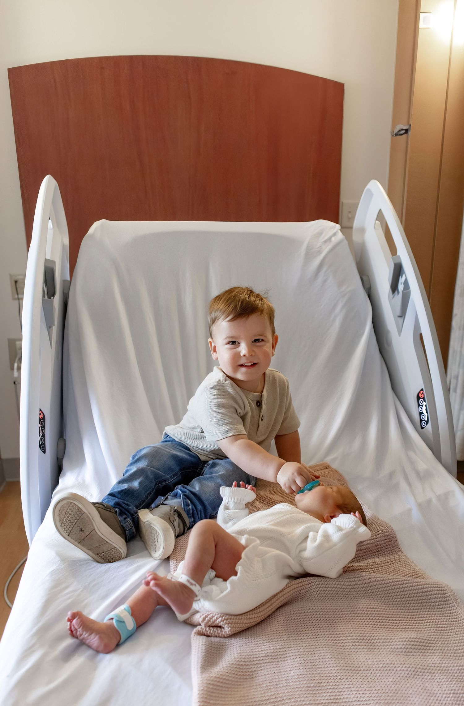 Two young children sit together on a hospital bed with white linens and wooden headboard in a medical setting.