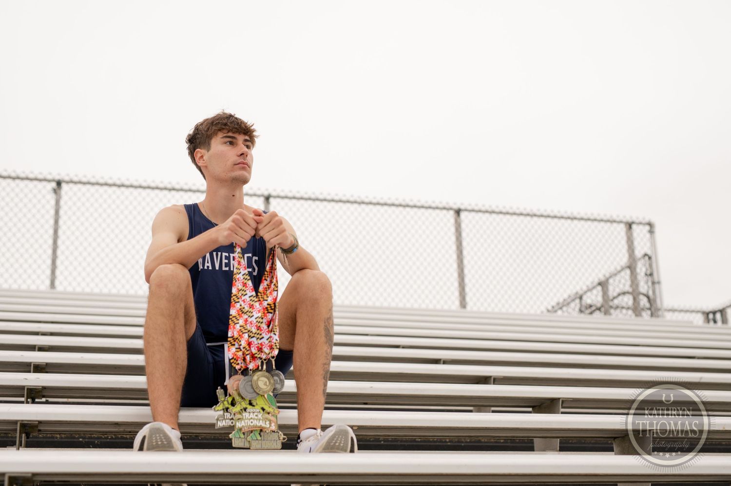 Athlete sits on metal bleachers wearing navy uniform and medals at track field.