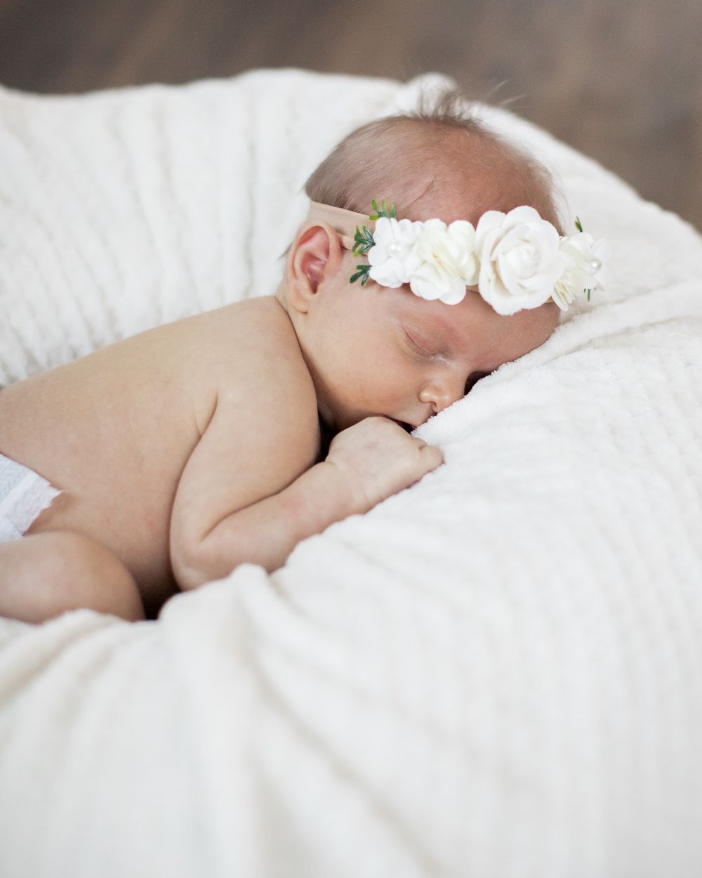 Sleeping baby wearing delicate white floral headband rests peacefully on white blanket.