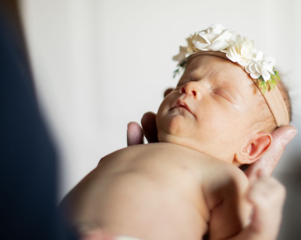 Sleeping newborn baby wearing delicate white floral headband against light background.