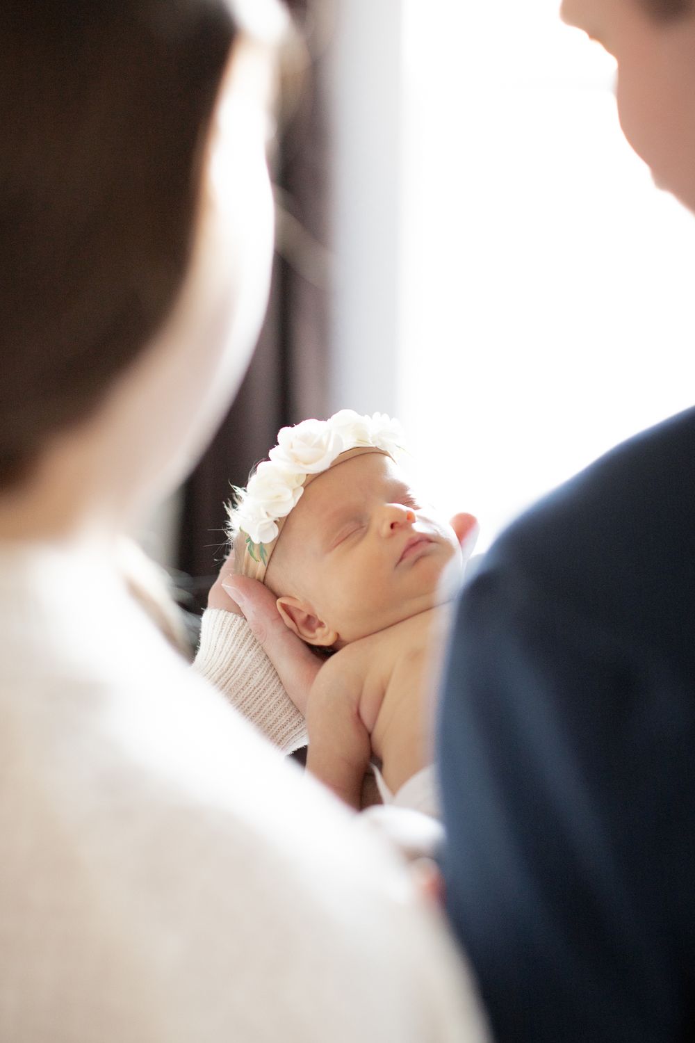 A sleeping newborn baby wearing a delicate white floral headband is cradled between two people.