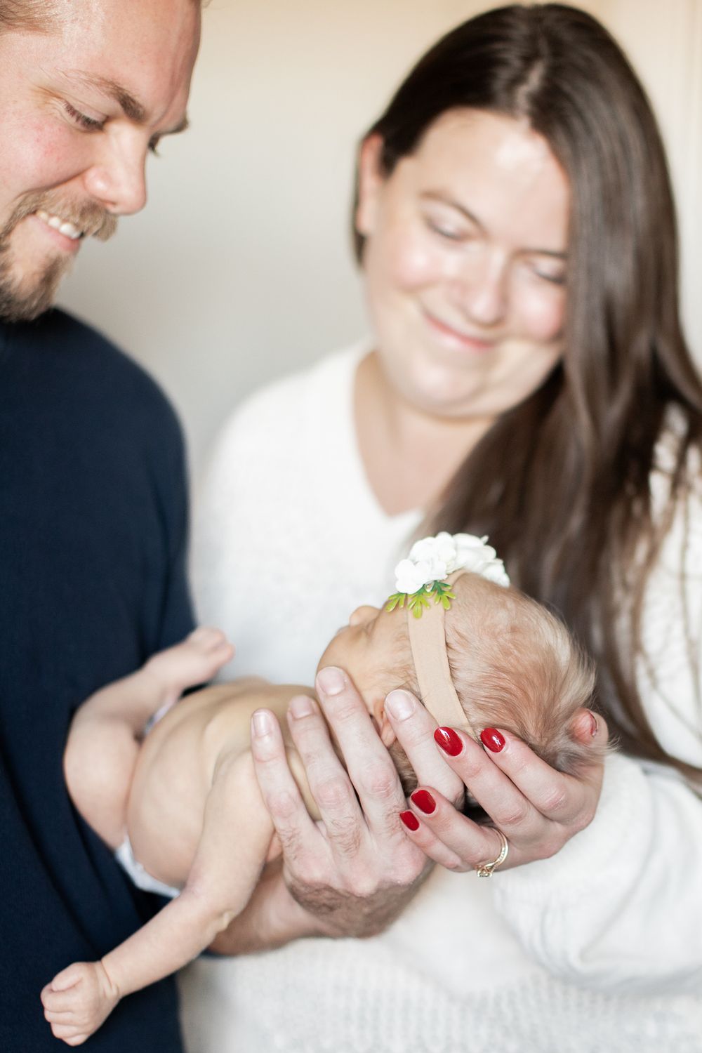 Parents in white and navy sweaters lovingly hold their newborn baby wearing a delicate white flower headband.