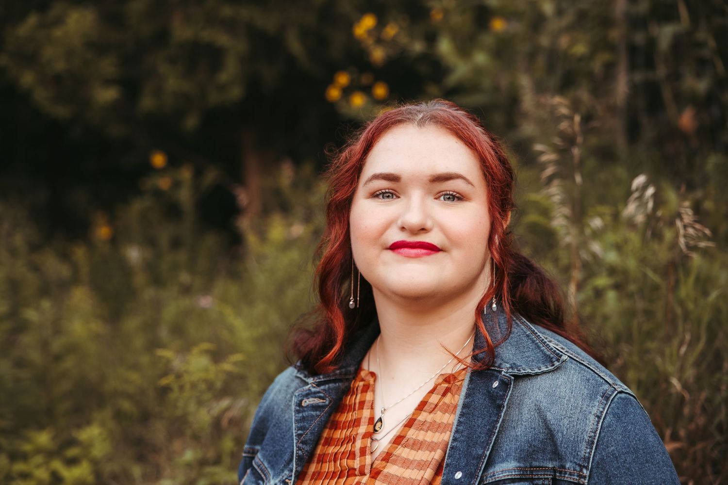 Senior wearing  jacket and orange plaid dress with autumn foliage in background. Late Summer Golden Hour Senior Photography in Ann Arbor