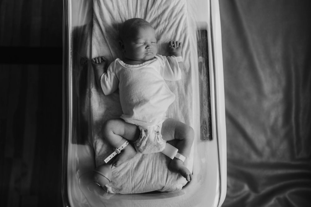 Black and white overhead shot of newborn baby lying on white bedding.