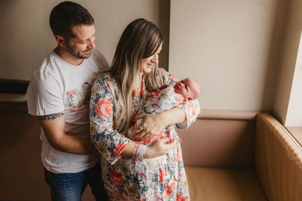 Parents holding their newborn baby in a tender family portrait by window light.