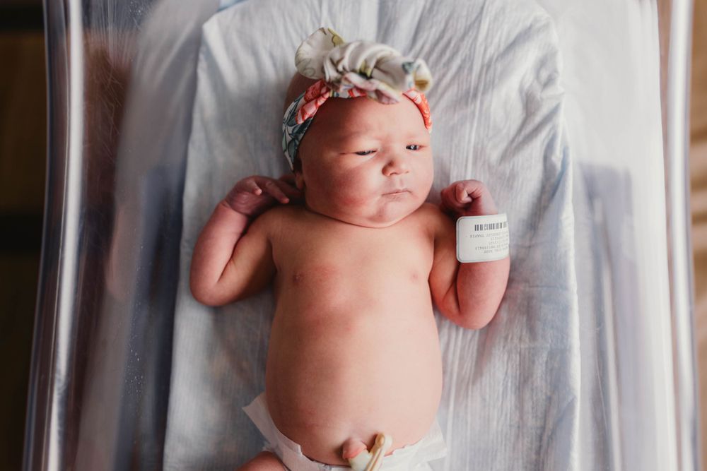 Newborn baby wearing floral headband lying on white hospital bassinet with medical monitoring device.