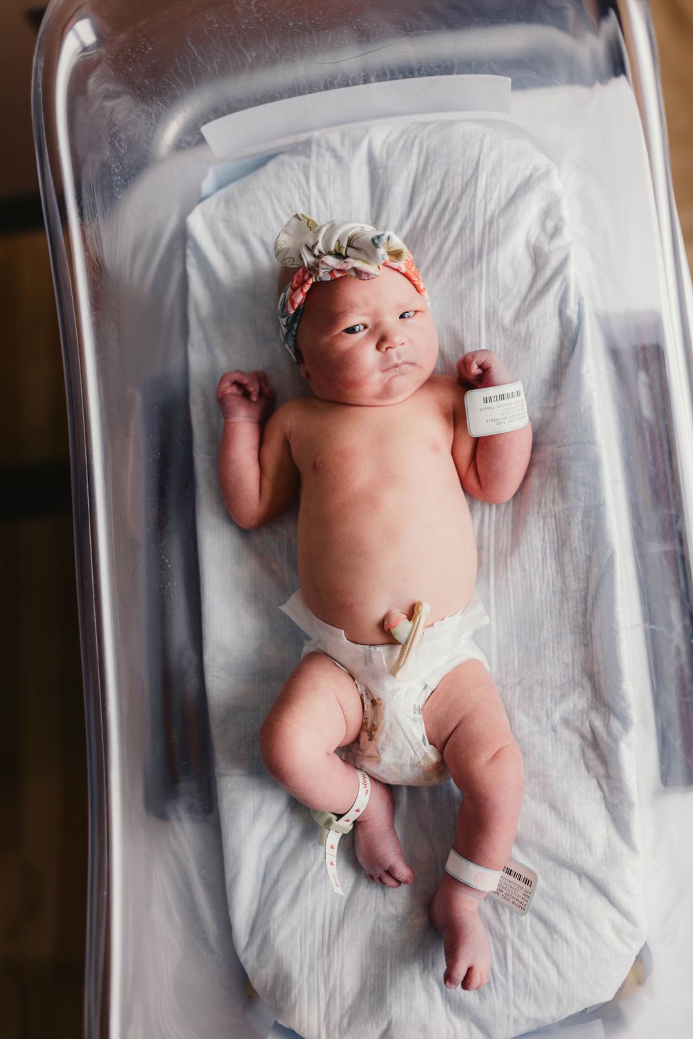 Newborn in diaper and floral headband resting in hospital bassinet with monitoring equipment.