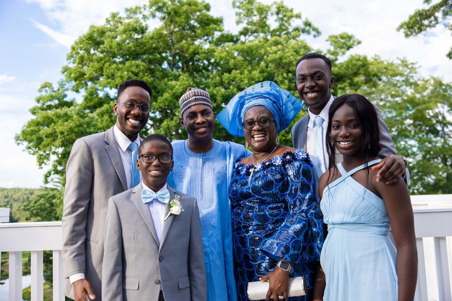 Family group poses outdoors in formal attire on a sunny day with green trees in the background.
