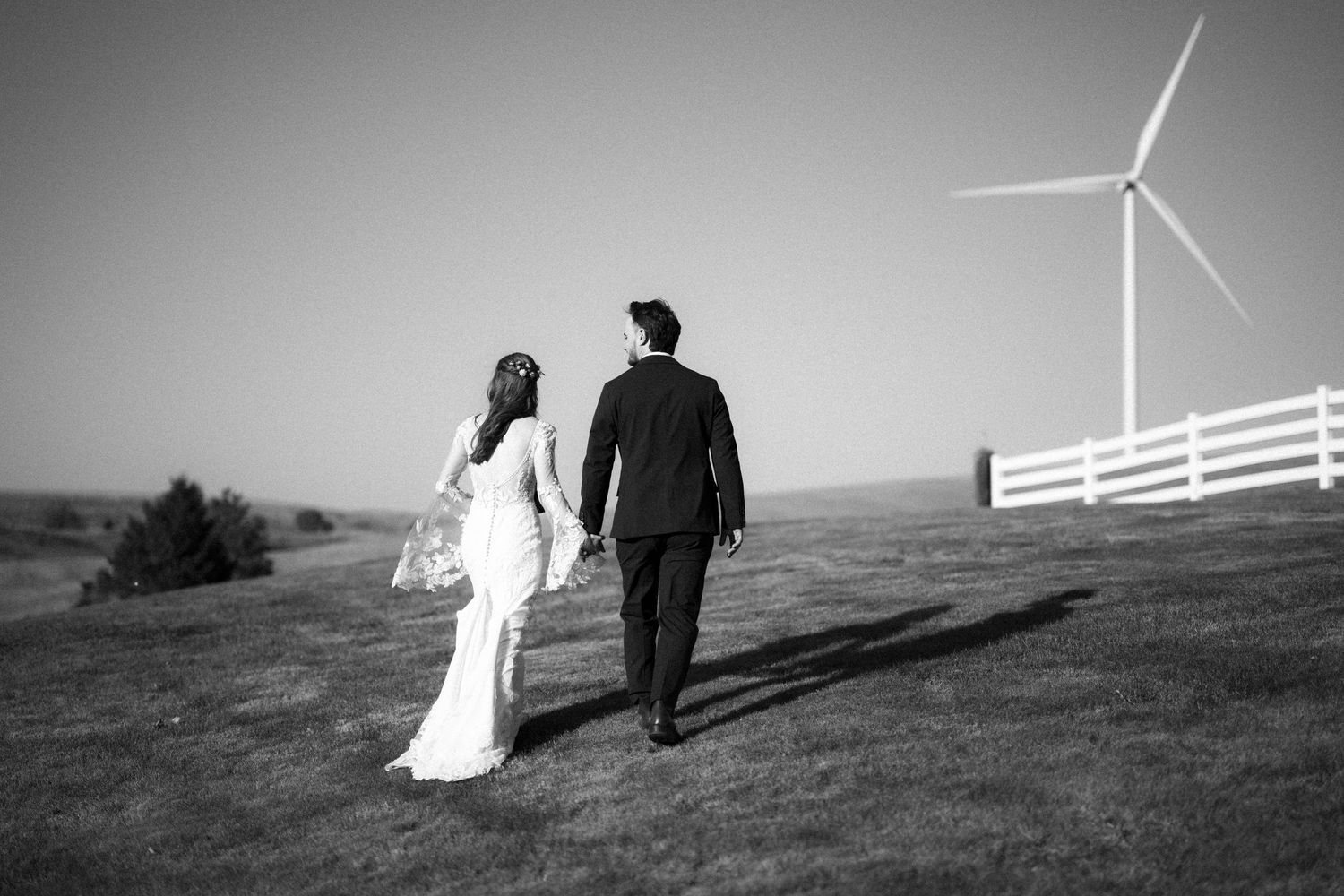Black and white image of couple walking on grassy hill with wind turbine in background.