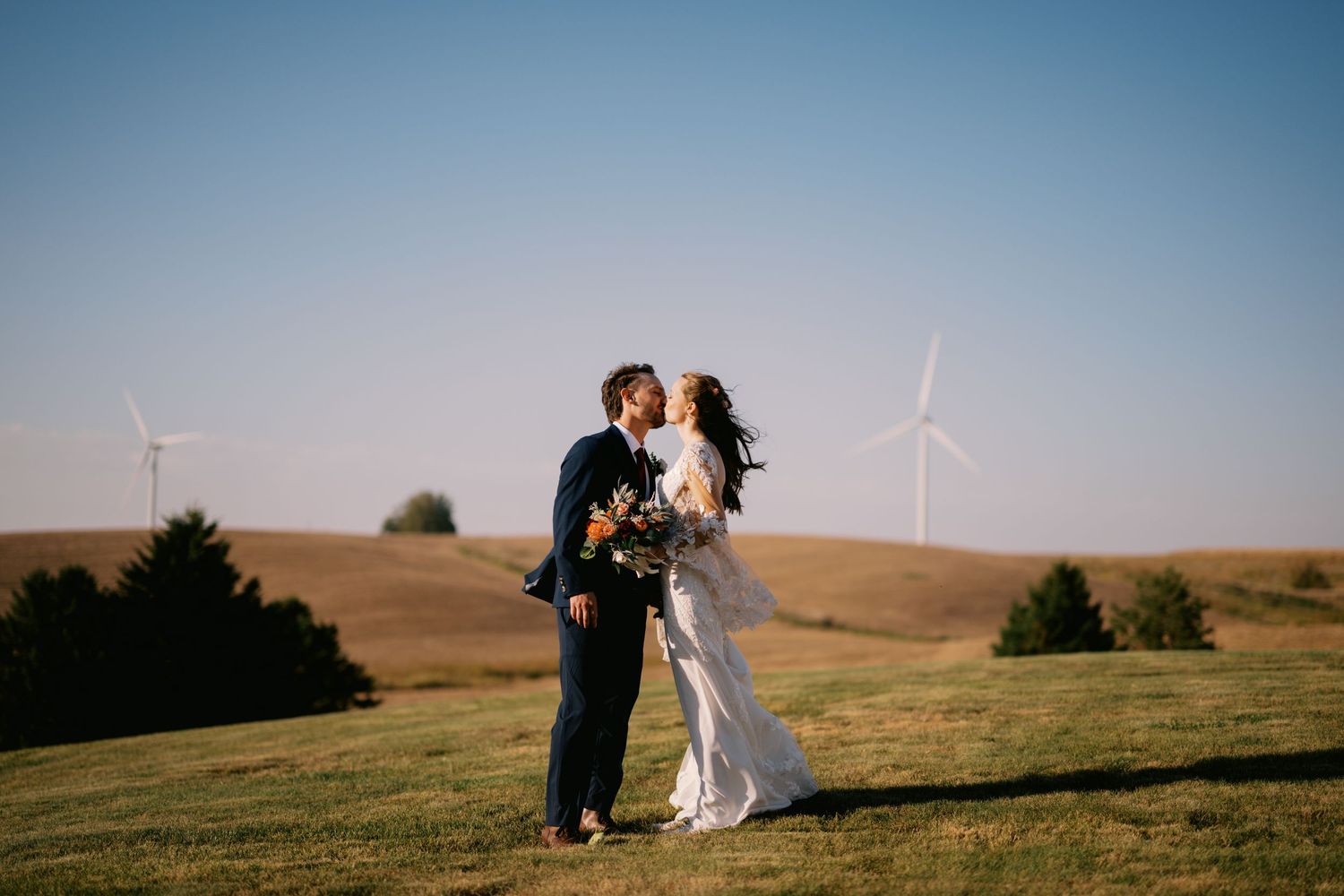 A couple in wedding attire embraces on a hilltop with wind turbines in the background during golden hour.