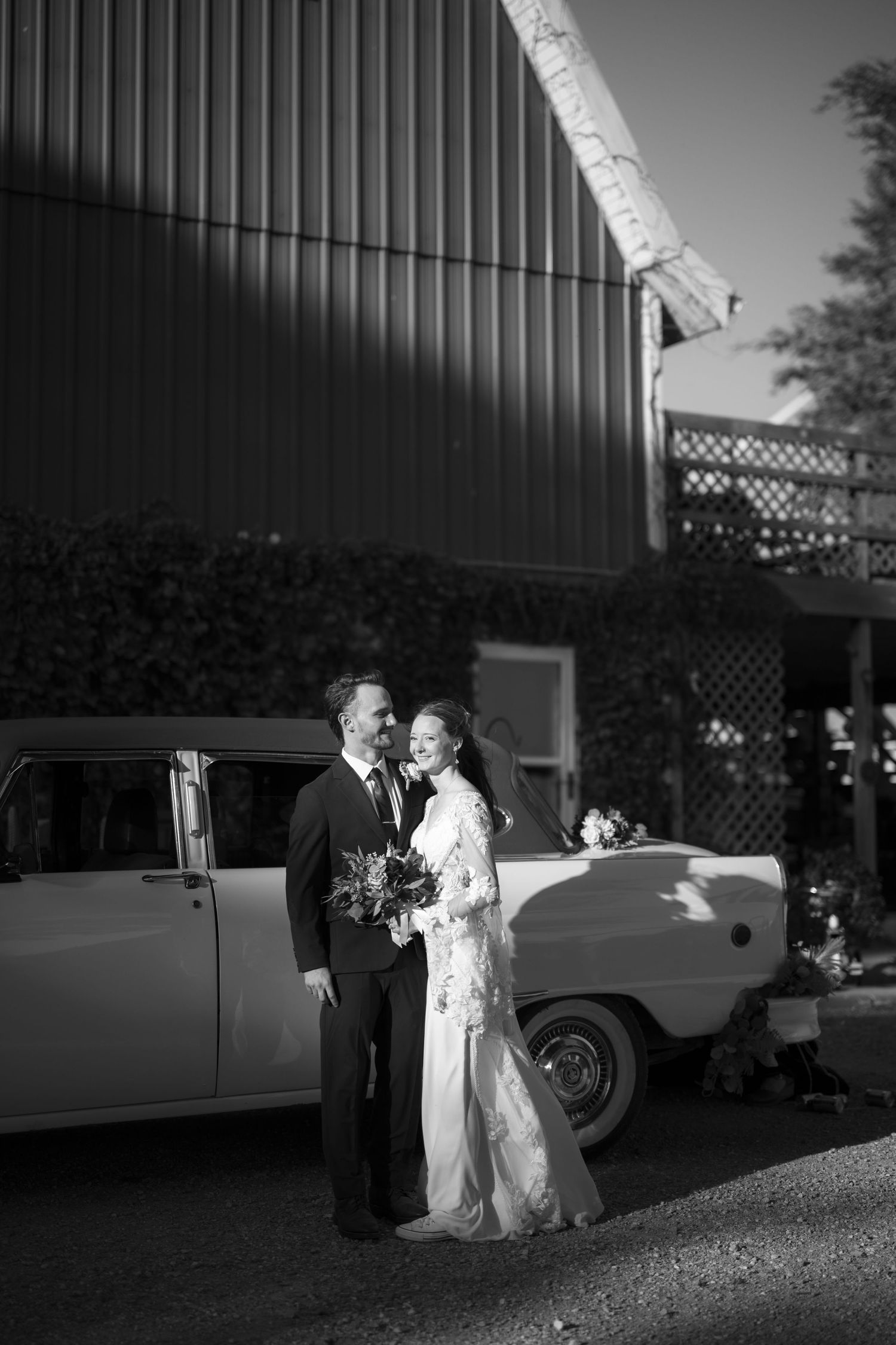 A couple stands beside a vintage car in black and white wedding photography with classic retro styling.