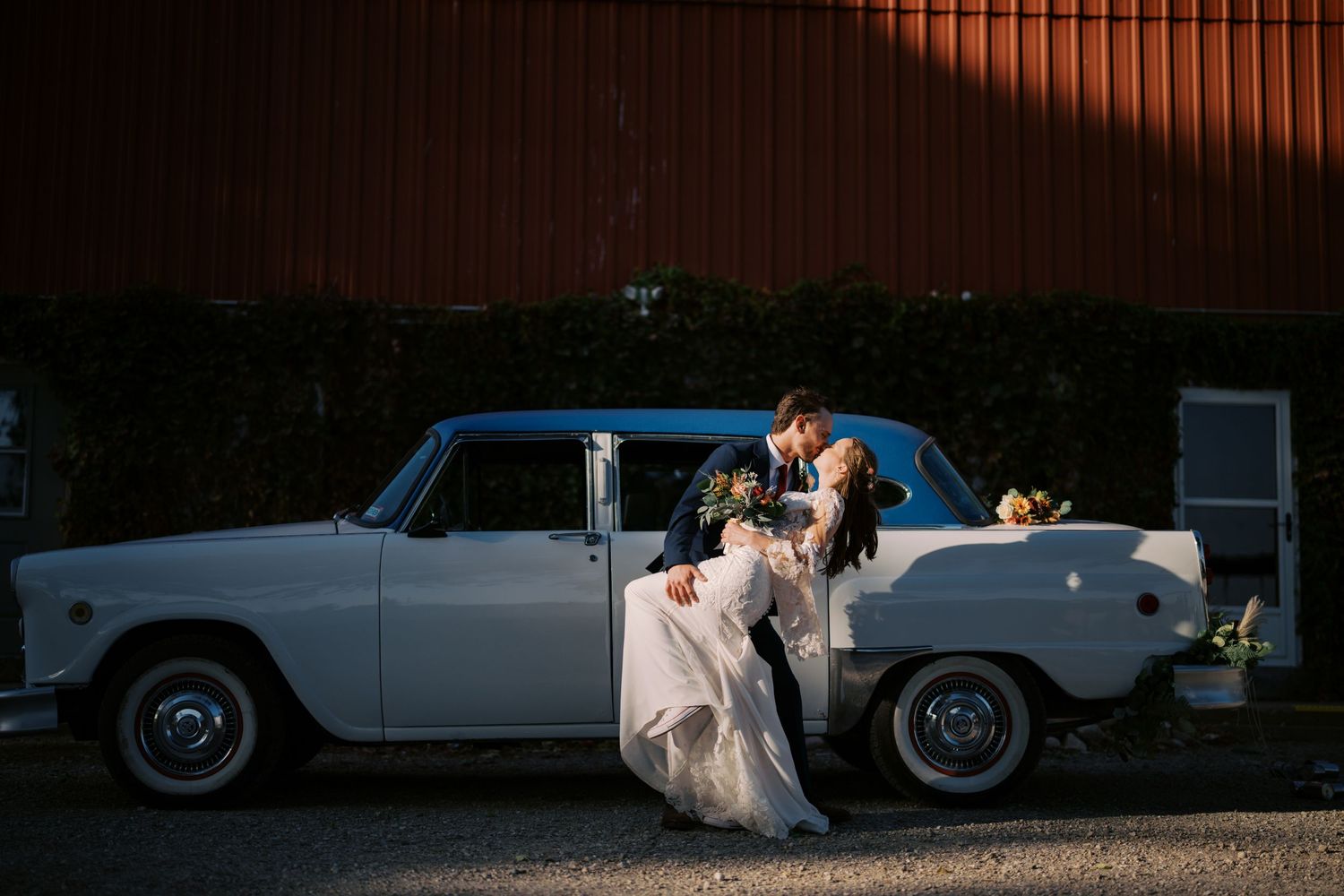 An elegant couple shares a romantic moment next to a classic vintage white car at sunset.