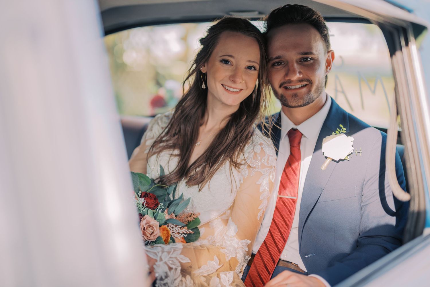 A couple poses together inside a vintage car on their wedding day wearing formal attire and holding a floral bouquet.