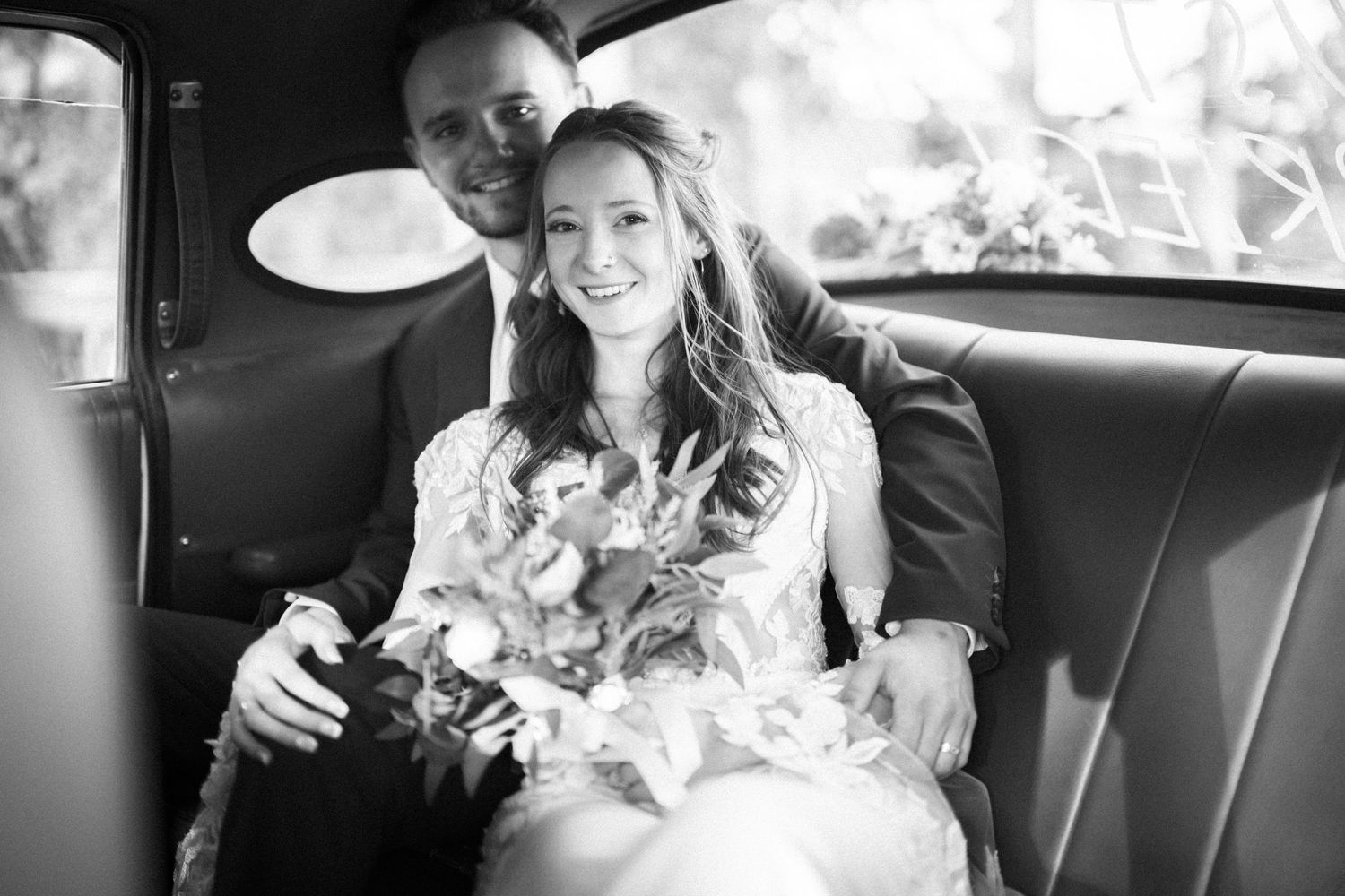 A bride and groom smile while sitting together in the back of a vintage car during their wedding celebration.