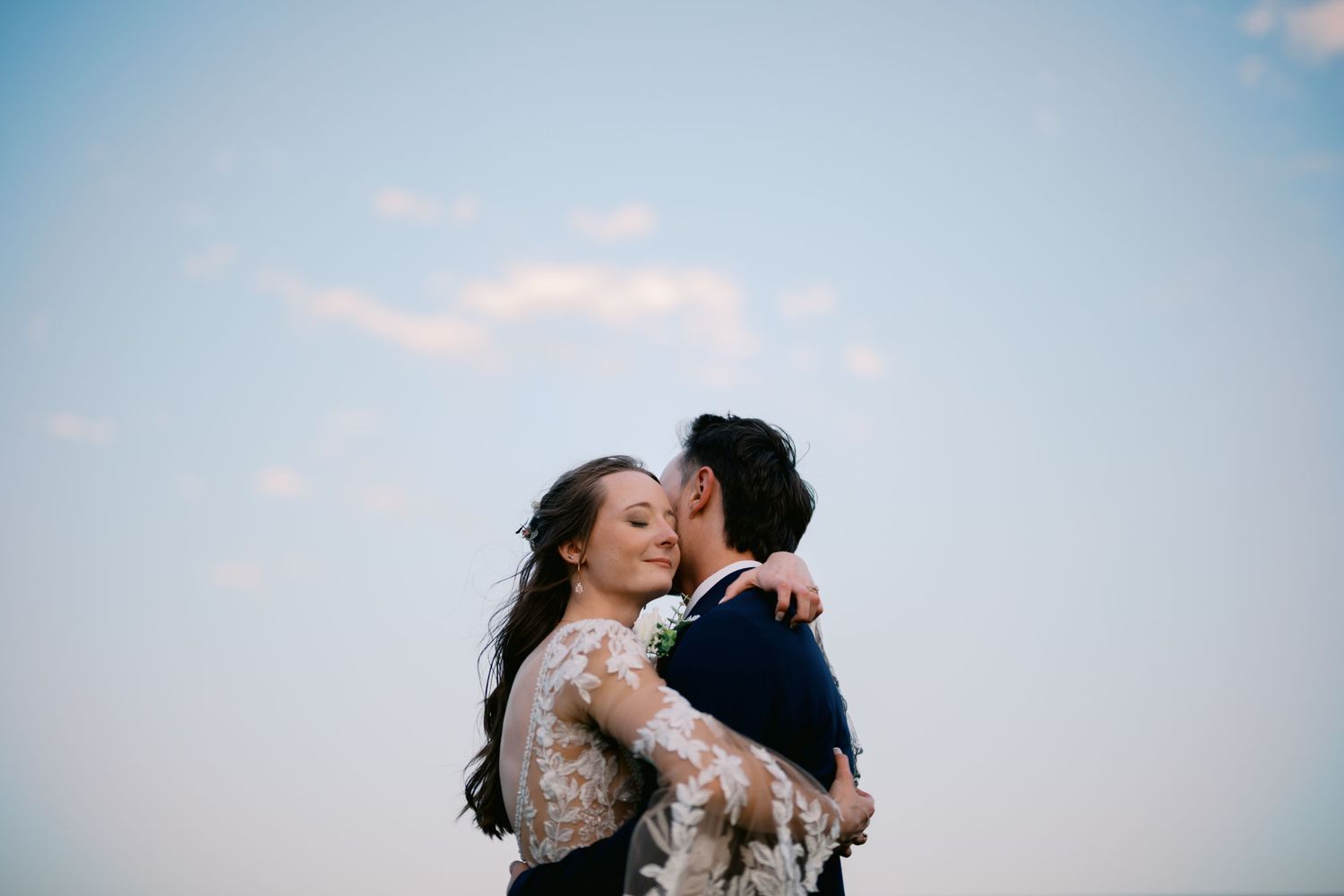 A couple embraces during a romantic wedding moment against a light blue sky.