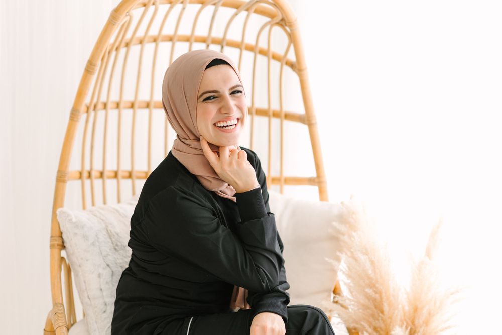 Smiling person in hijab sitting on rattan chair with white cushions and pampas grass decor.