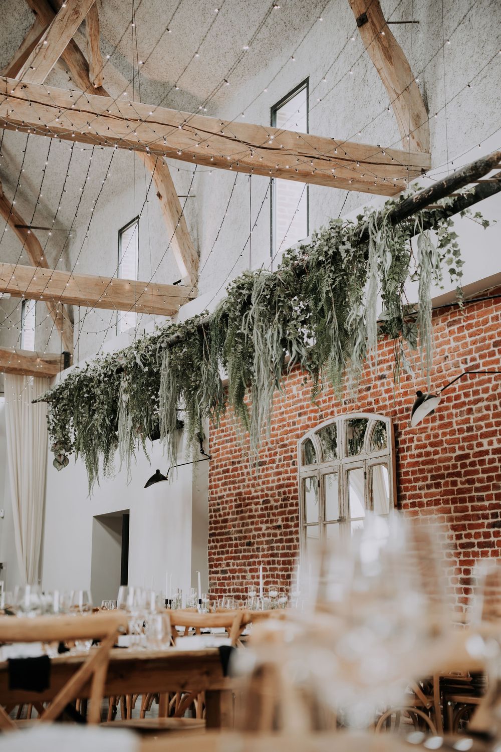 Rustic barn wedding reception with hanging greenery garland, exposed wooden beams, brick wall, and elegantly set dining tables.