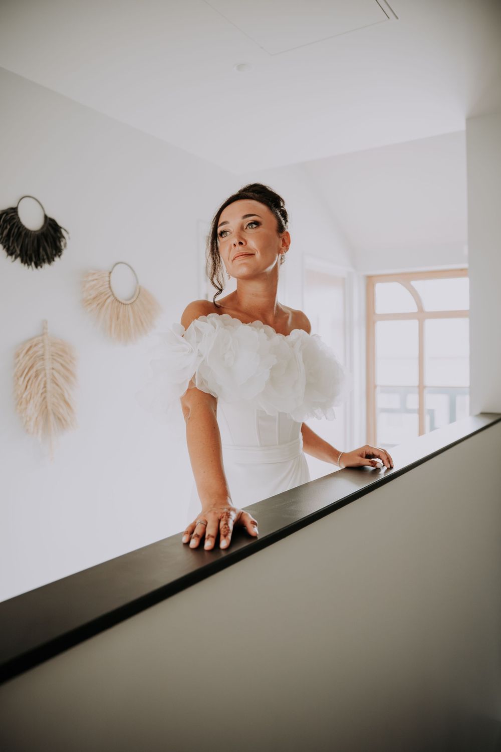 Bride in white off-shoulder ruffle gown poses gracefully on staircase with boho wall decor in bright modern interior.