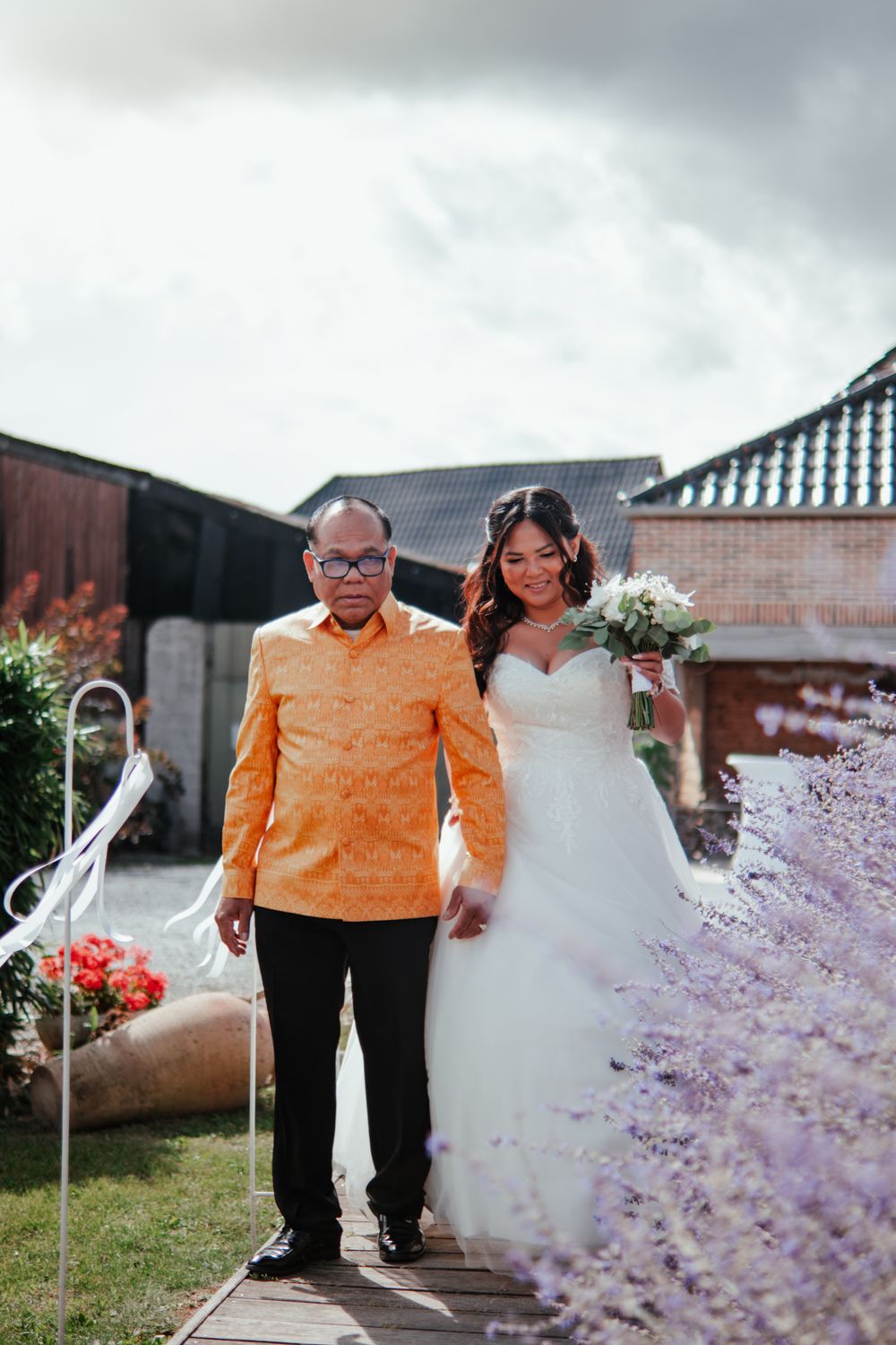 A couple walks together through a garden path with lavender flowers, wearing an orange shirt and white dress.