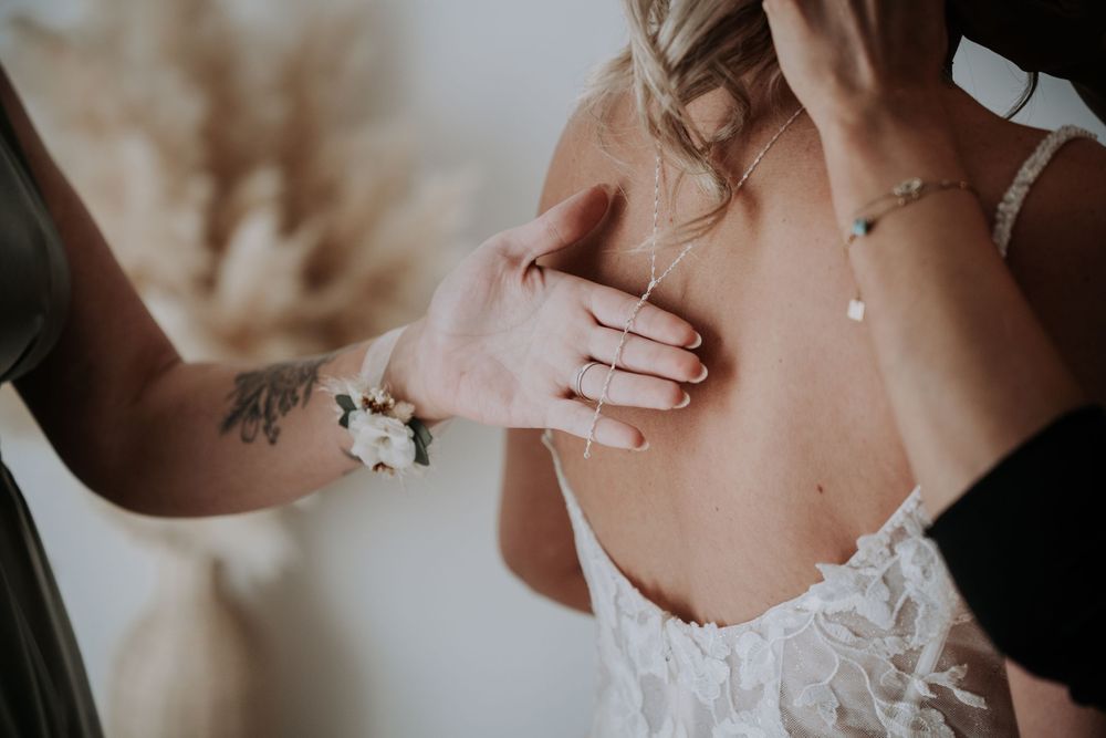 Tattooed bridesmaid fastening the back of a lace wedding dress on a bride wearing delicate jewelry.