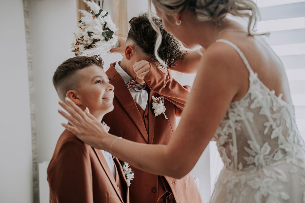 Bride in lace gown helps young boy in rust suit while groom stands nearby during wedding preparations.