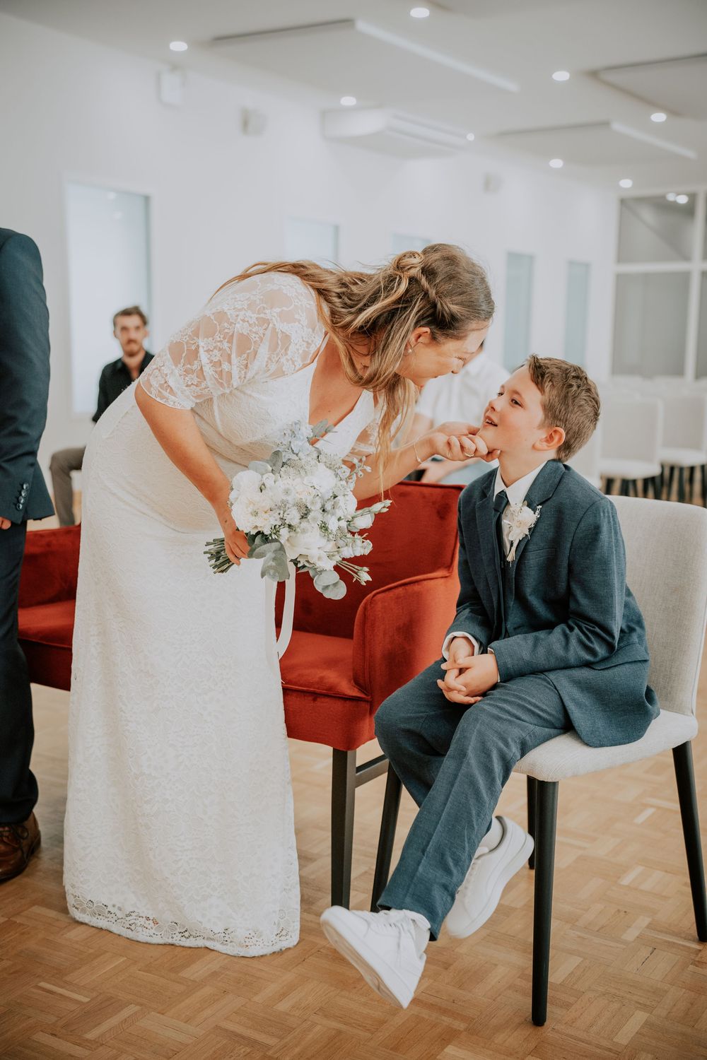 Bride in lace gown holding white bouquet leans toward smiling boy in navy suit at wedding ceremony.