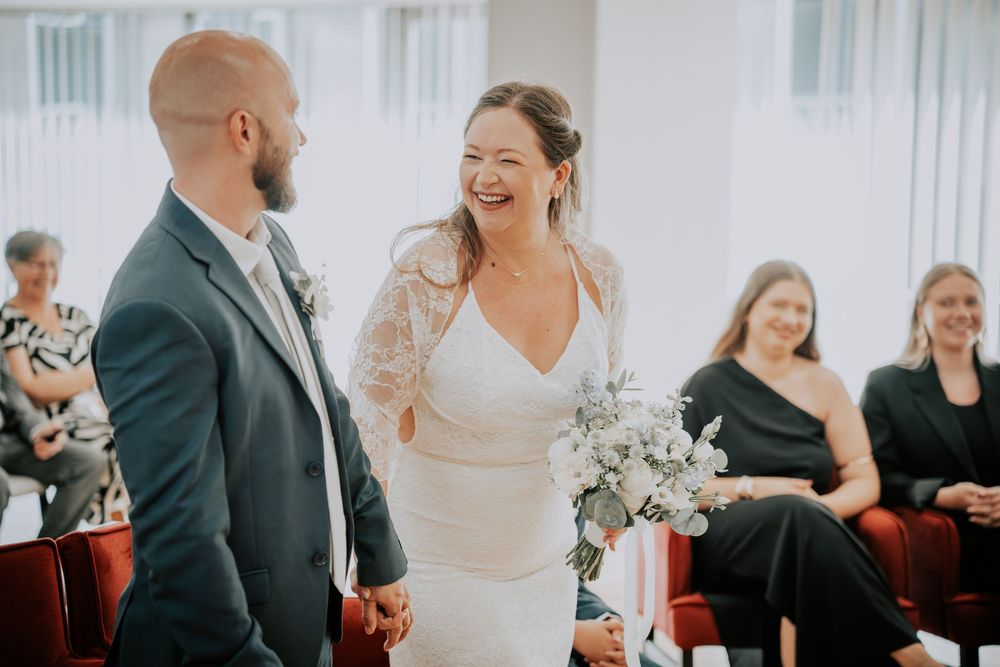 Bride laughing joyfully with groom during intimate wedding ceremony, holding white floral bouquet, guests seated in background.