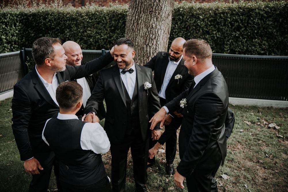 Groom in black tuxedo with bow tie laughing with groomsmen outdoors near a tree at a wedding celebration.