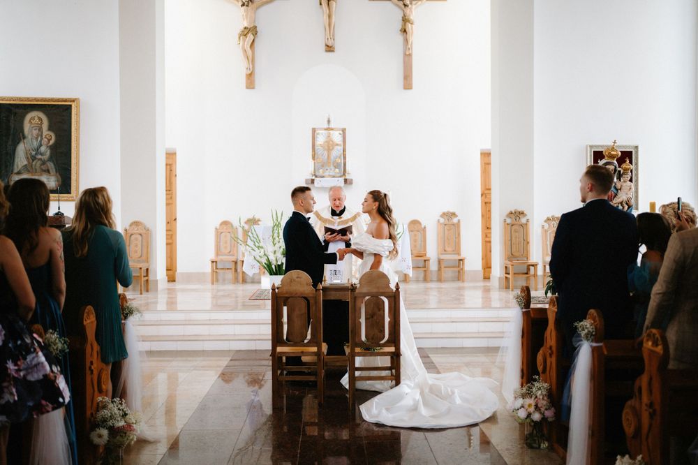 Wedding ceremony taking place in a bright white church with wooden accents and religious symbols.