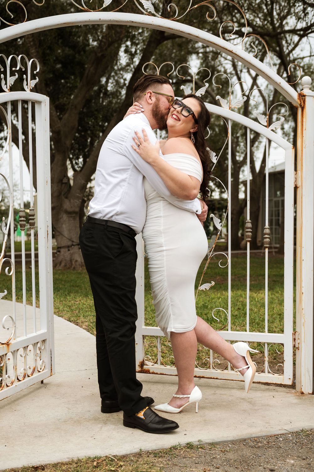 Playful romantic moment between couple under ornate white metal archway in garden setting.