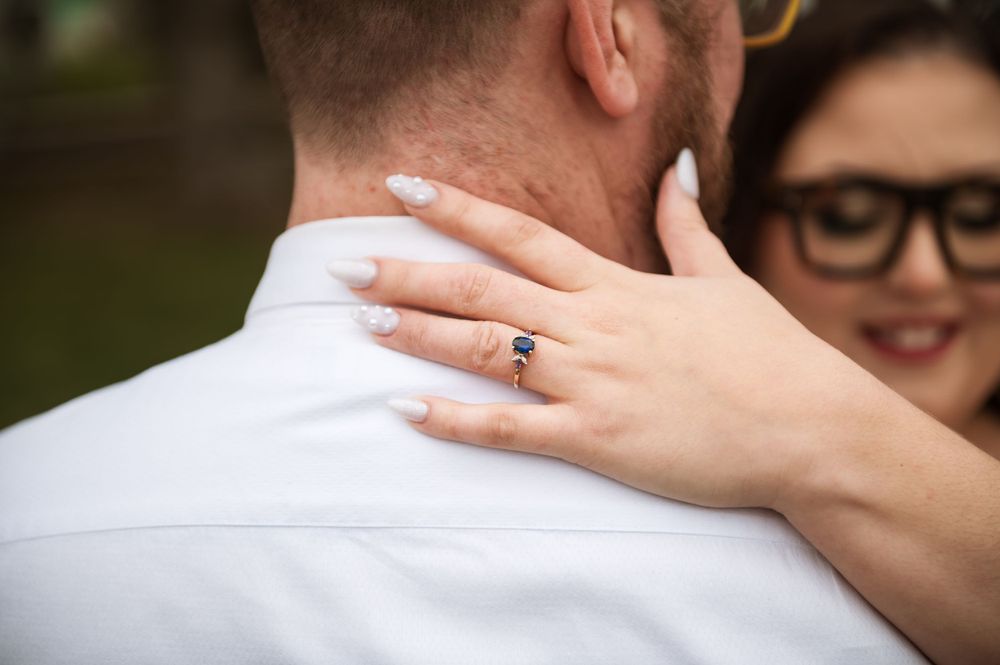 Close-up shot of a hand with an engagement ring resting on a white-shirted shoulder.