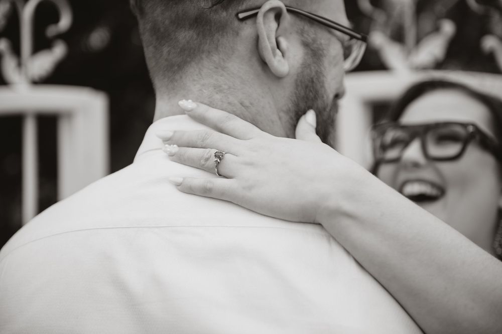 Black and white close-up of a hand with an engagement ring touching a shoulder.