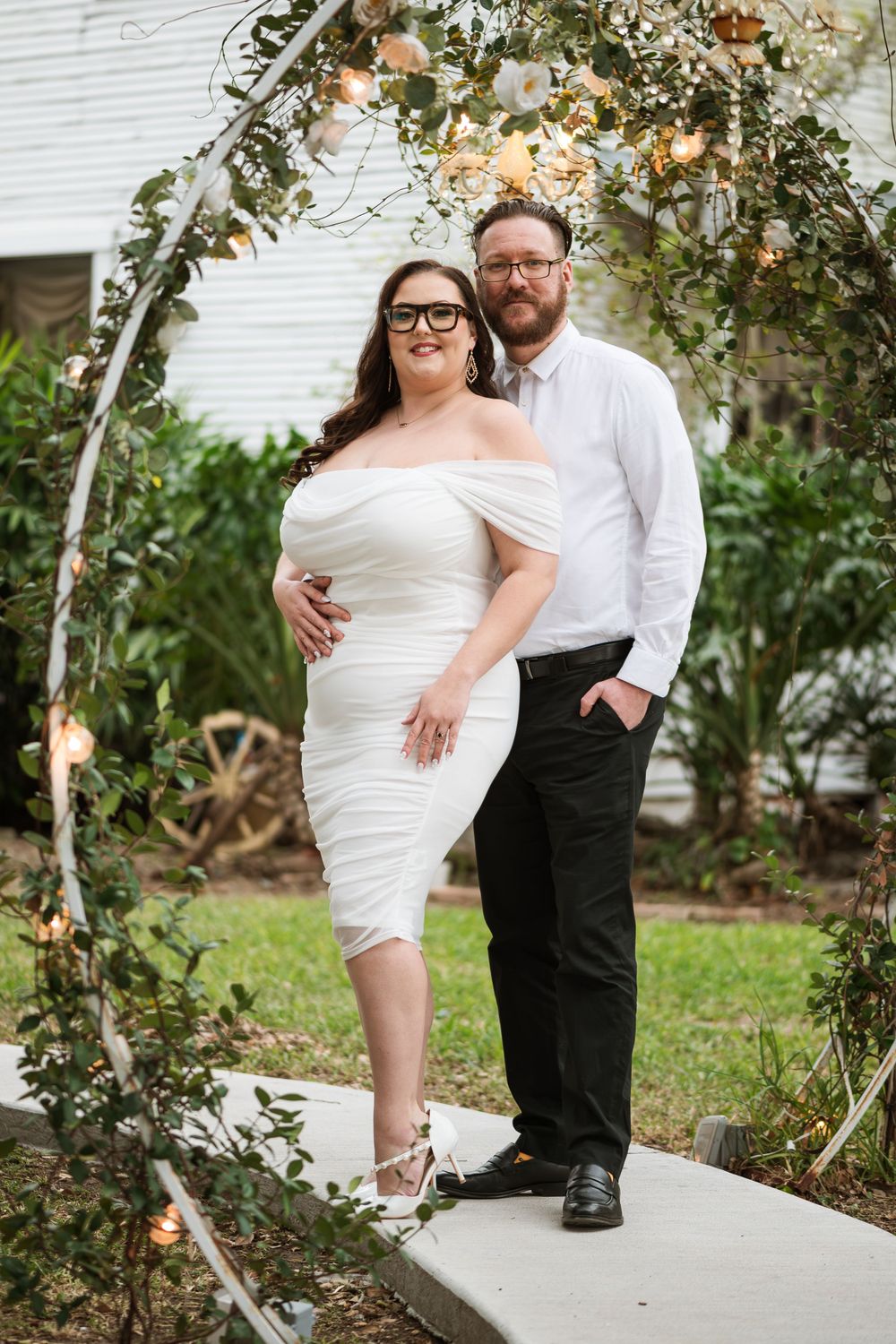A couple poses under a floral arch in formal attire during their wedding photoshoot.
