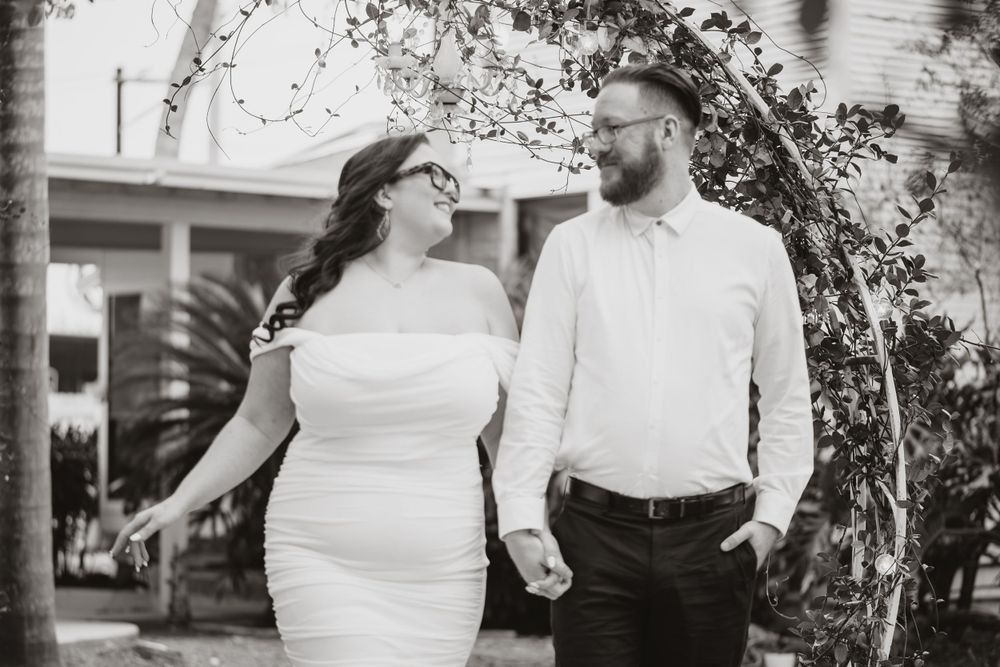 A black and white portrait of a couple in wedding attire holding hands under trees.