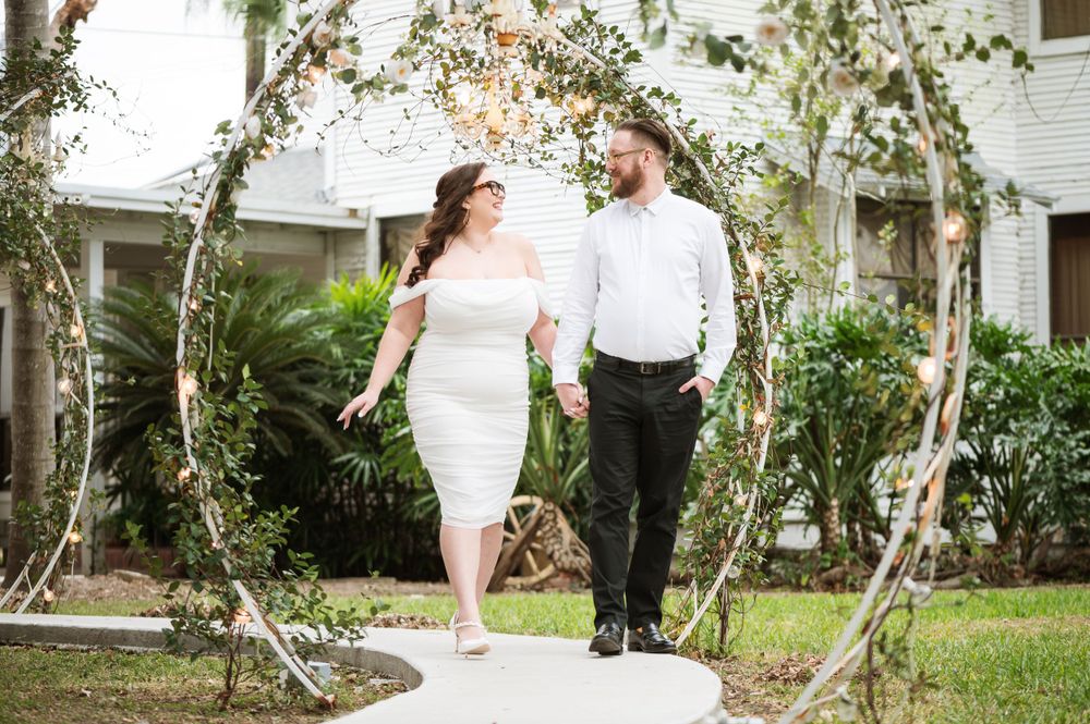 Couple walking along garden path under floral arch in wedding attire.