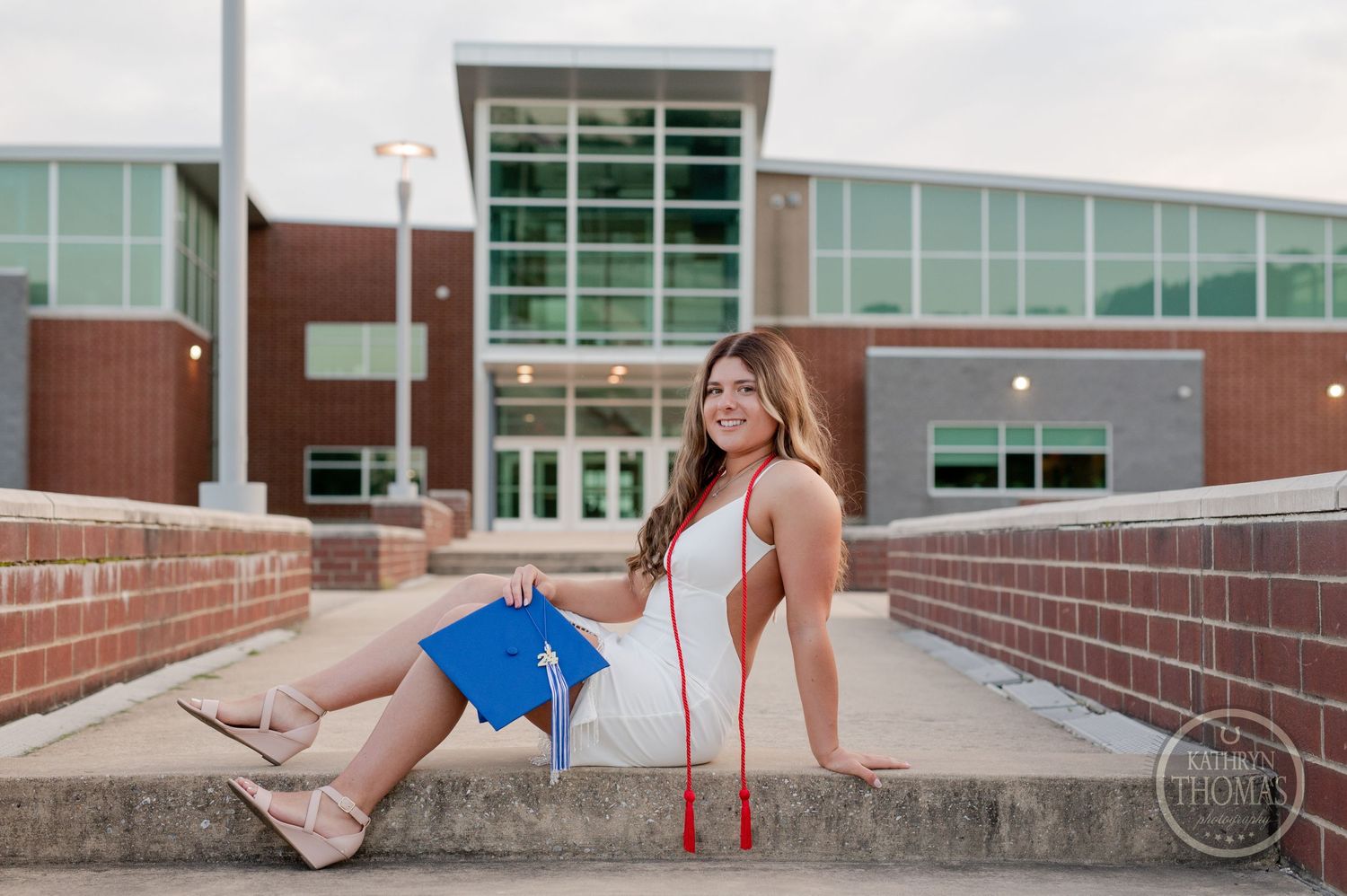 Graduate in white dress sits on brick wall ledge holding blue graduation cap.