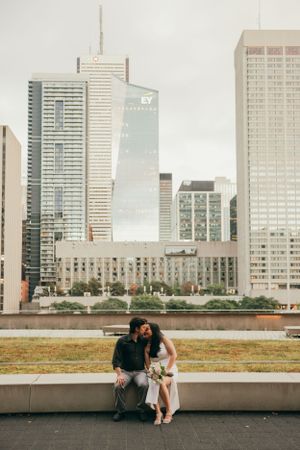 A couple sits on a bench with Toronto's downtown skyscrapers in the background.