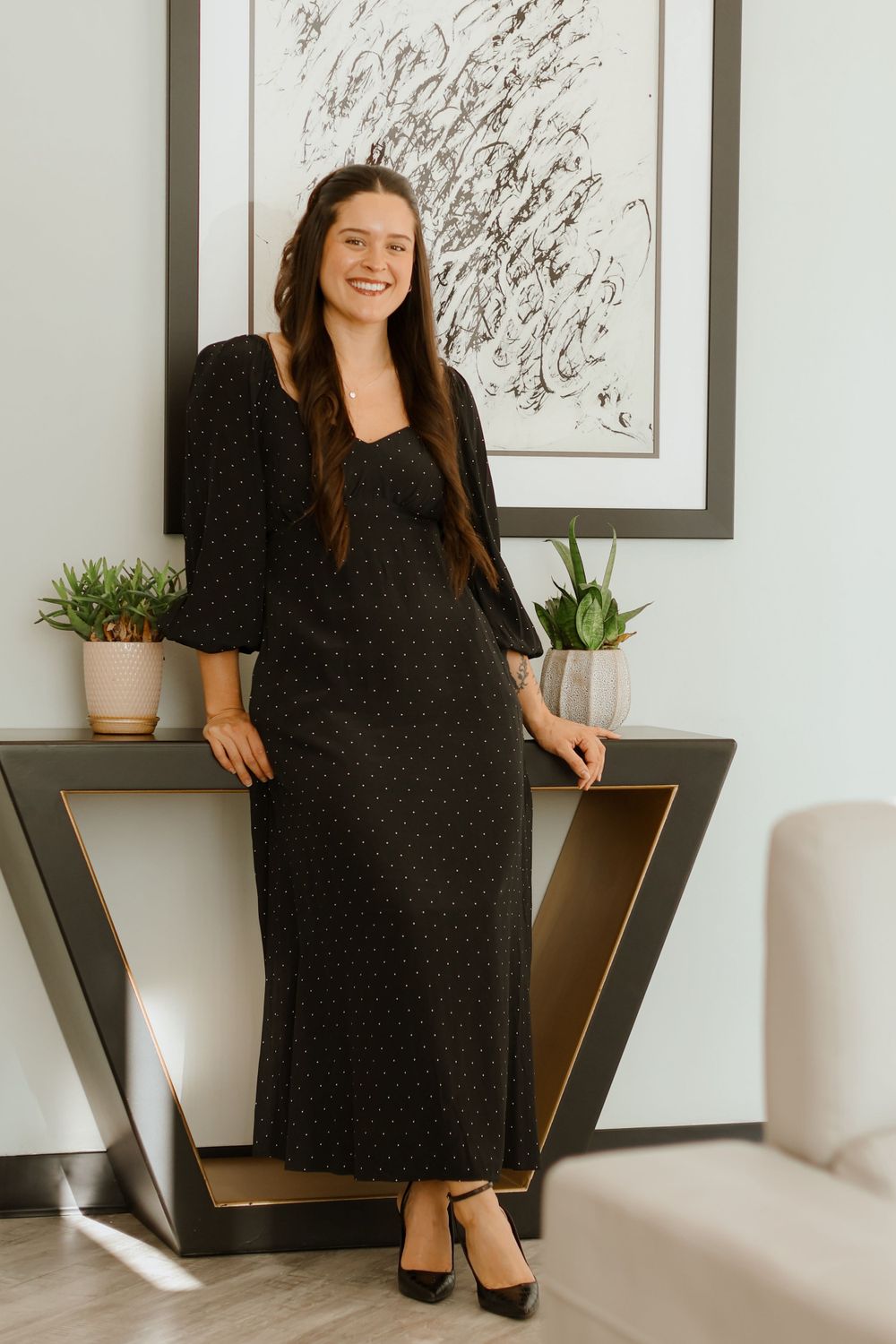 Person in black dress seated on chair with artwork and plants in background.