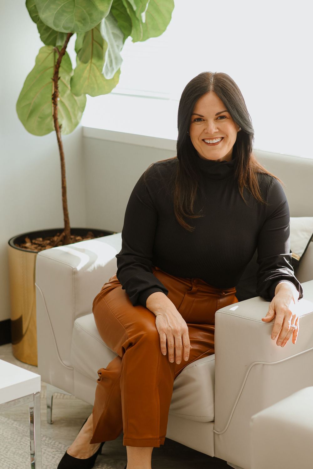 Woman in black top and brown pants relaxing on white armchair beside indoor plant.