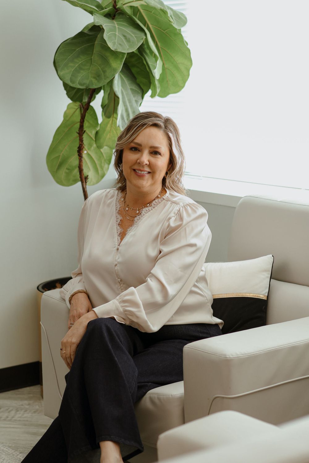 Professional portraits in cream blouse against white wall with fiddle leaf fig plant.