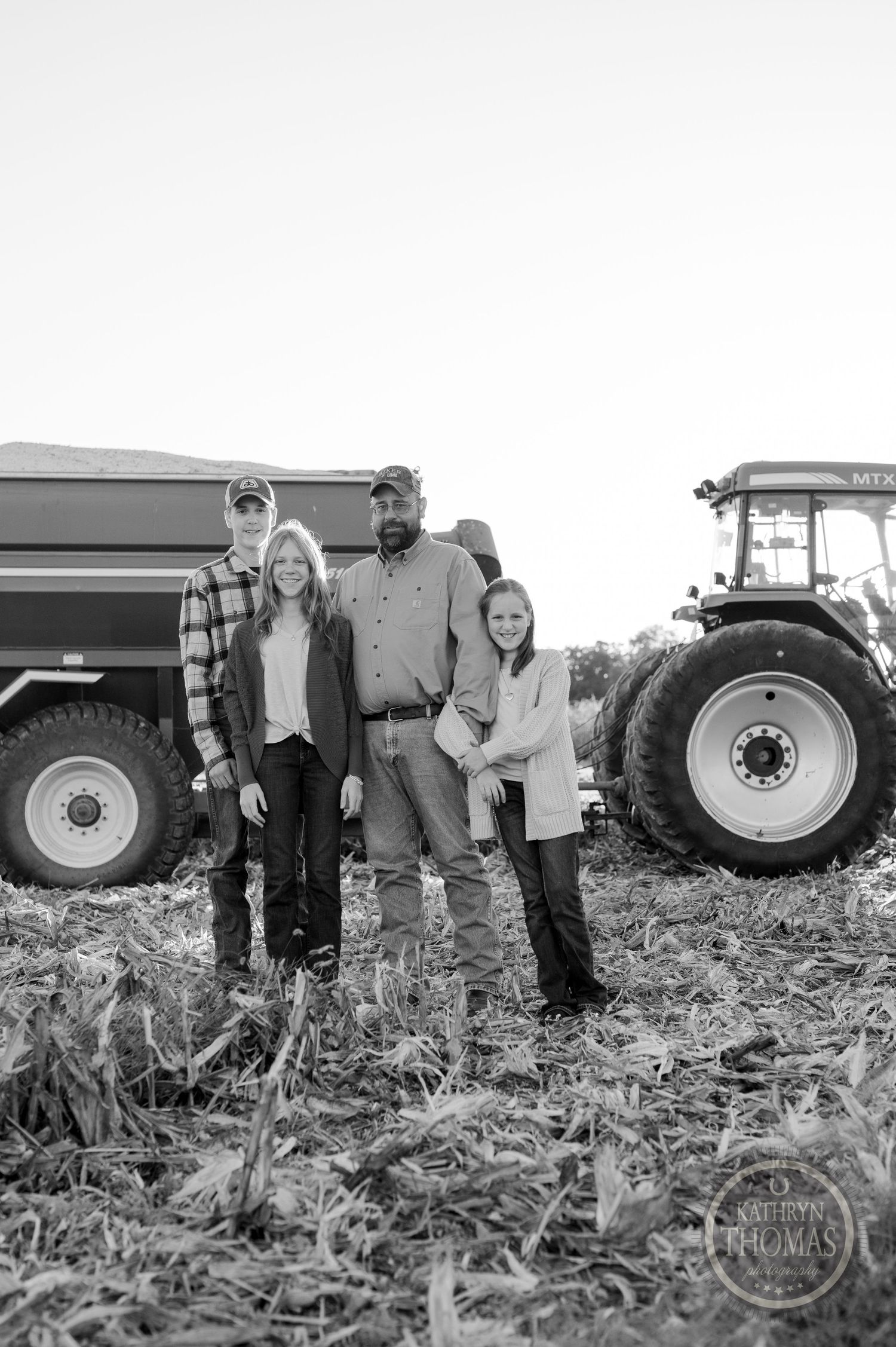 A farming family poses together in front of their equipment on agricultural land in black and white.