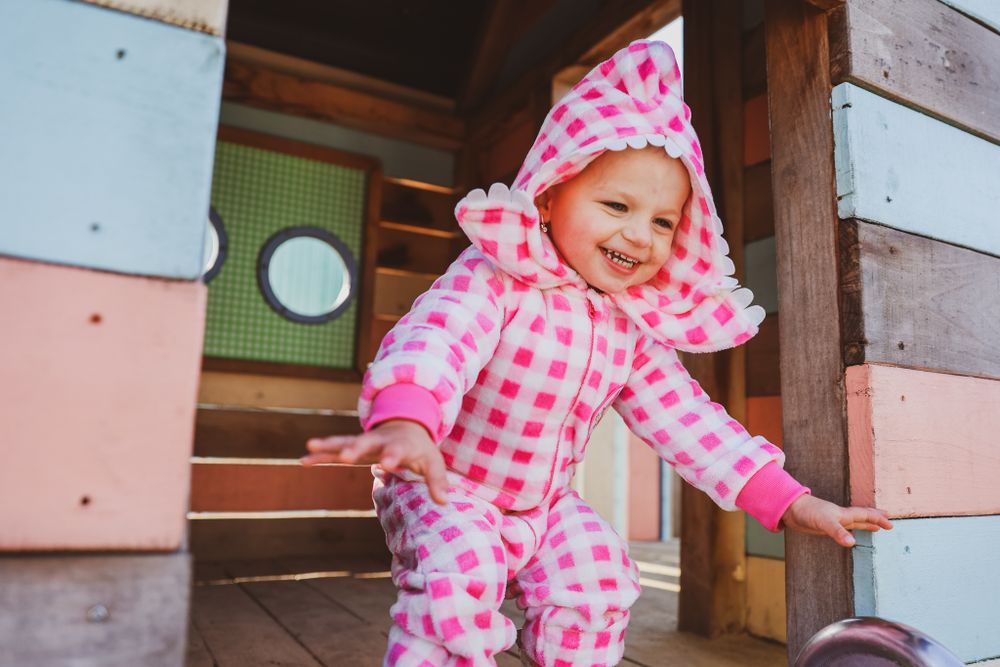 Child wearing pink and white checkered onesie playing in colorful playhouse.