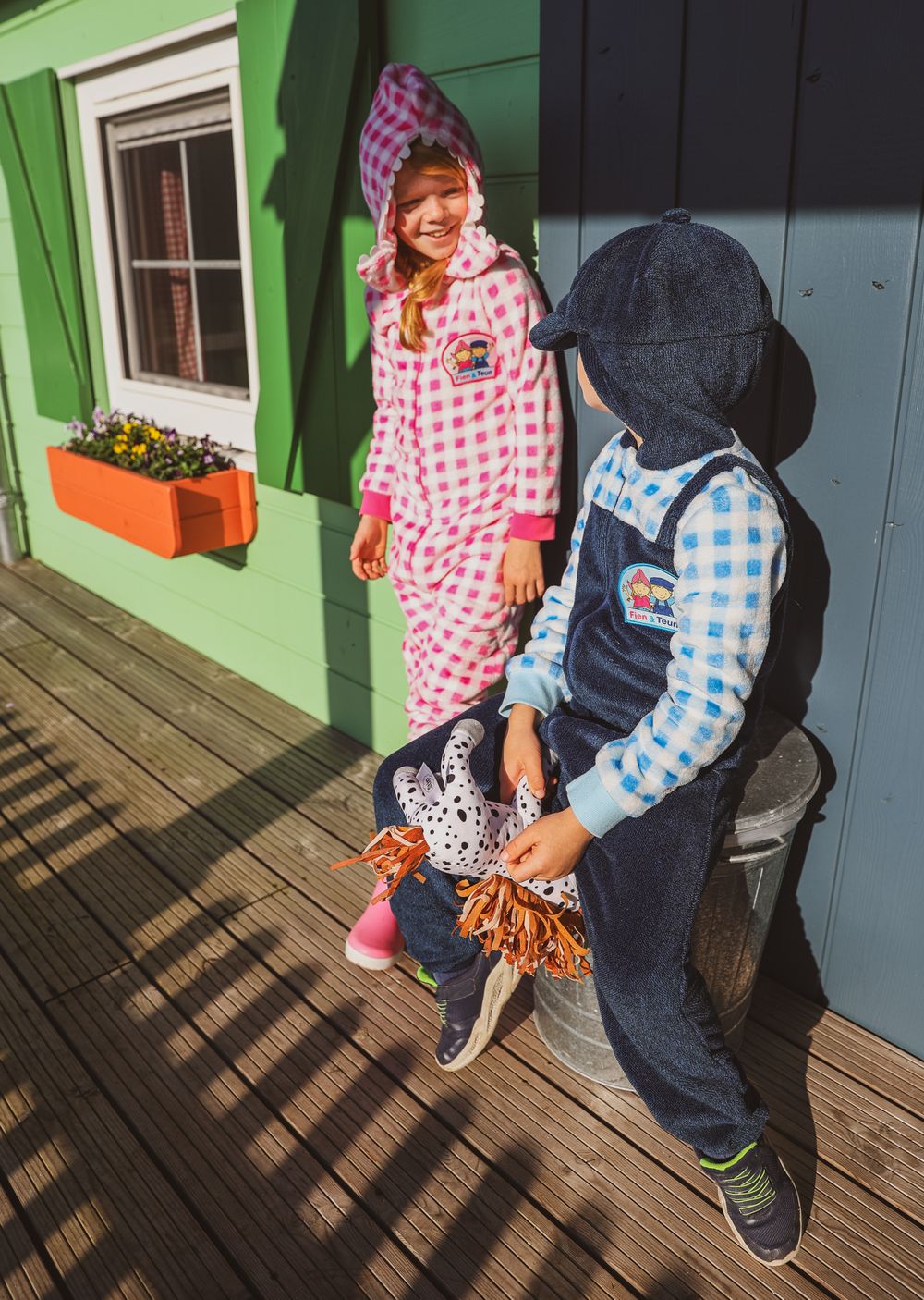 Two children wearing matching gingham pattern sleepwear posing on wooden porch with green house exterior.