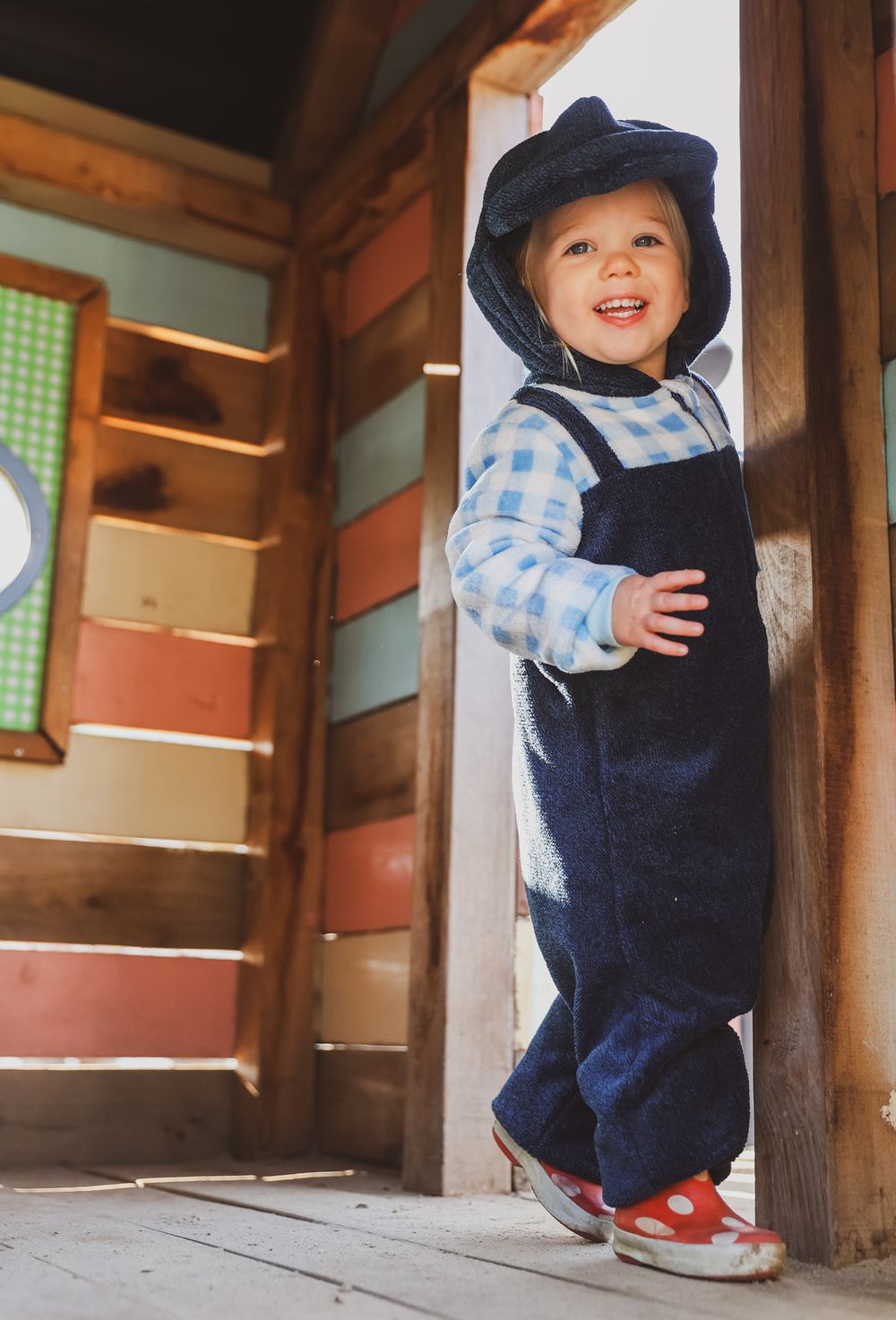 Small child in navy overalls and checkered shirt standing in wooden doorway.