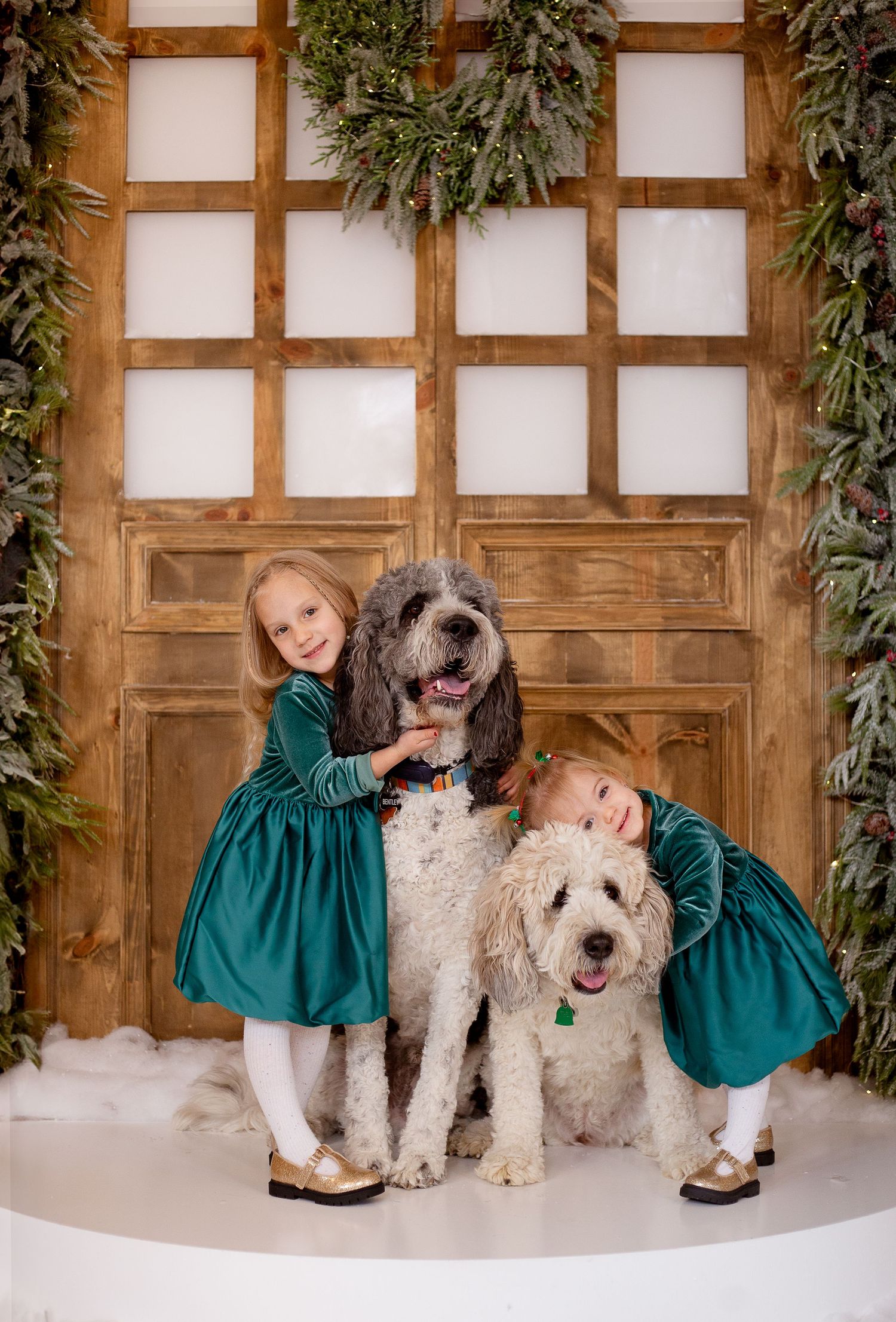 Two children in matching green dresses pose with two large white shaggy dogs in front of a wooden door with holiday decor.