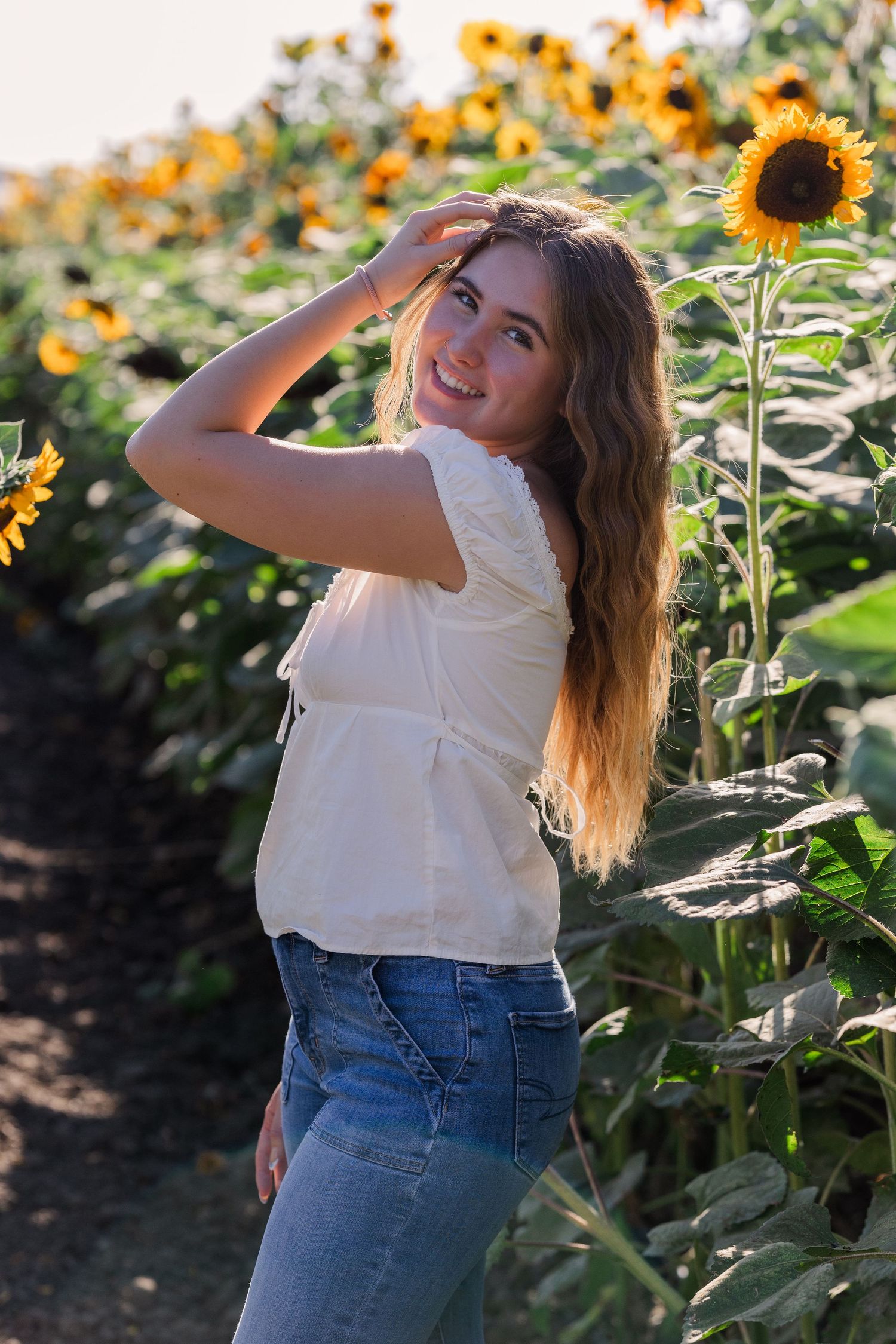 Person in white top and jeans posing near blooming sunflowers in garden.