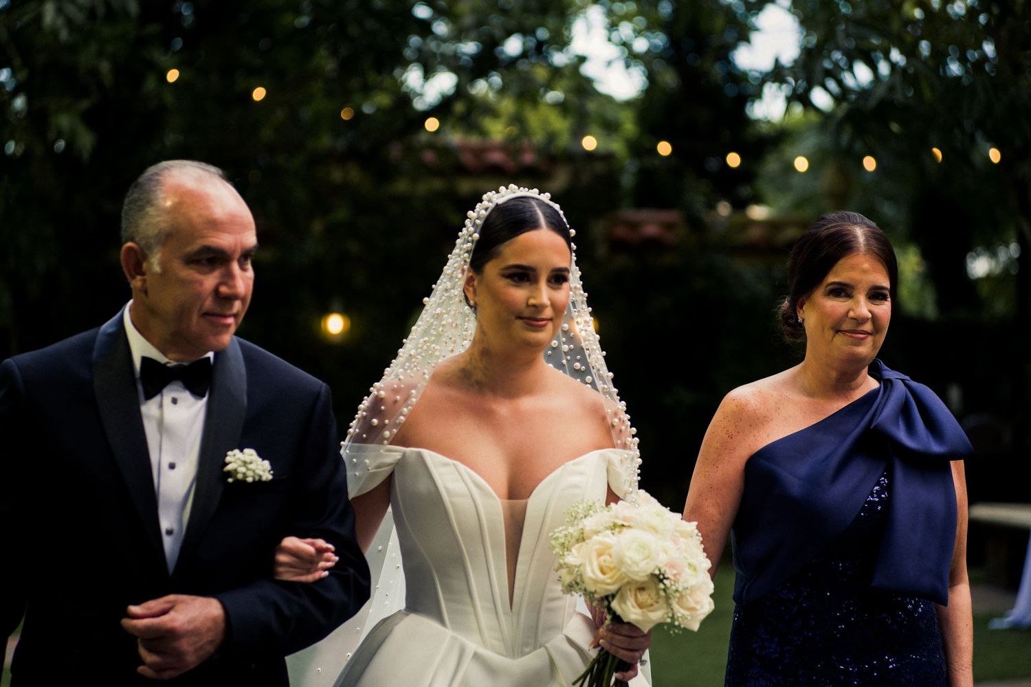 An elegant wedding party walking outdoors under string lights at dusk.